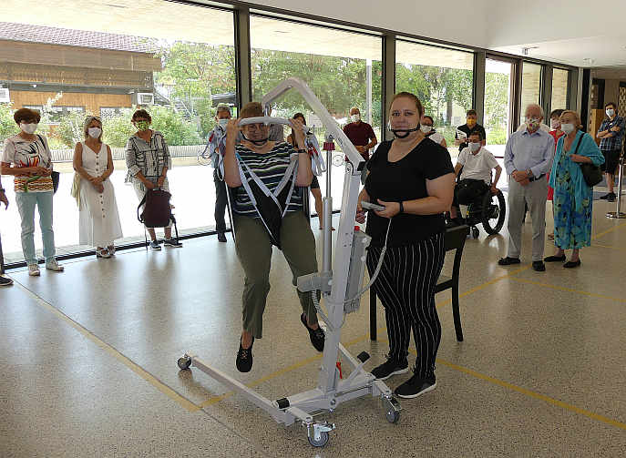 ...Nina Stockinger hebt Jutta Pagel-Steidl (hilfsweise von einem Stuhl) mit dem Hebetuch „per Knopfdruck“ mit dem Lifter in die Höhe...<br />Foto: © Mara Sander