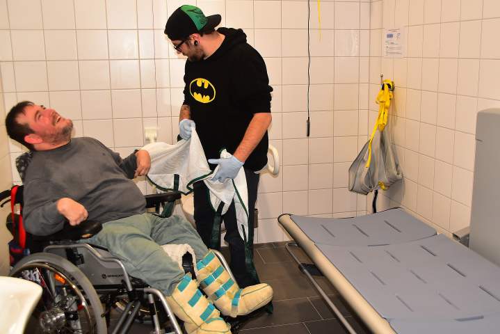 Assistent Fabian Fürst platziert das Hebetuch so, dass Inklusionsbotschafter Peter Maier mit dem Hebelifter problemlos auf die Pflegeliege schweben kann ...<br />Foto: © Mara Sander