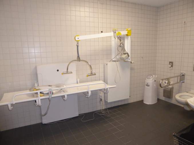 Die »Toilette für alle« ist eine Rollstuhl-Toilette, die zusätzlich mit einem höhenverstellbaren Wandklappliege, Lifter und luftdicht verschließbarem Windeleimer ausgestattet ist.