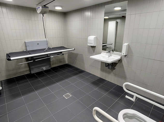 Die rund 10 qm großebarrierefreie Toilette ist zusätzlich ausgestattet mit höhenverstellaren Wandklappliege für Erwachsene (mit abklappbarem Seitengitter), einem Deckenlifter sowie einem luftdichten Windeleimer. Die „Toilette für alle“ ist mit Euro-Schlüssel rund um die Uhr zugänglich.