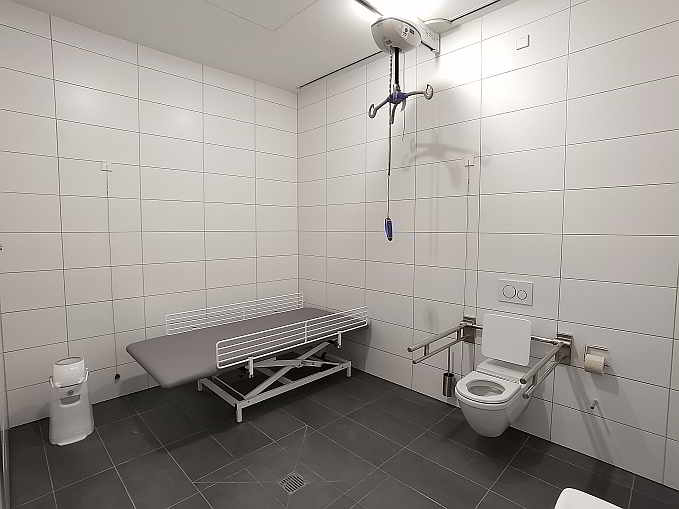 Die rund 11 qm große Rollstuhltoilette ist zusätzlich ausgestattet mit einer frei stehenden höhenverstellbaren Pflegeliege für Erwachsene, Deckenlifter und luftdichtem Windeleimer