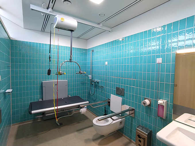 Die rund 8 qm große „Toilette für alle“  (einschl. Dusche) ist ausgestattet mit einer höhenverstellbaren Wandklappliege (Länge 140 cm) sowie einem Deckenlifter. Sie ist rund um die Uhr mit Euro-Schlüssel zugänglich.